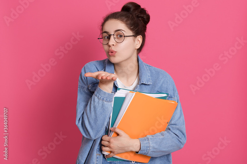 Lovely and tender college girl, student with knot, sending air kiss to camera, holding folders with documents, happy student wearing denim jacket and glasses.