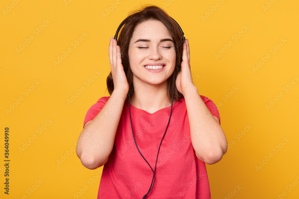 Young smiling brunette girl with closed eyes wearing red t-shirt, listening music via headphones, enjoying favorite songs and relaxing, isolated over yellow background. Young smiling brunette girl with closed eyes wearing red t-shirt, listening music via headphones, enjoying favorite songs and relaxing, isolated over yellow background.