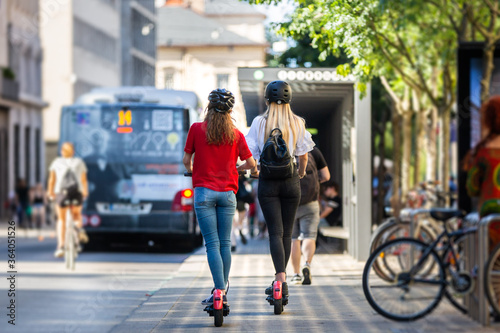 Rear view of trendy fashinable teenager girls riding public rental electric scooters in urban city environment. New eco-friendly modern public city transport in Ljubljana, Slovenia.