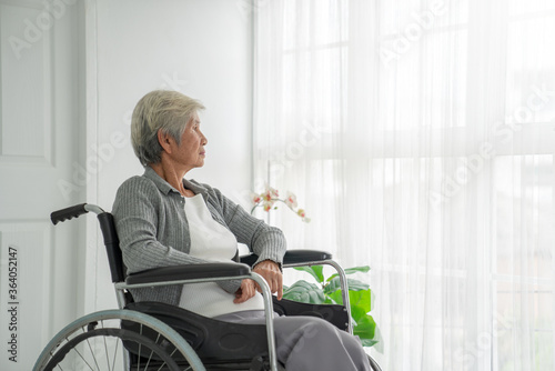 Rear view of a senior woman sitting on wheelchair looking outside the window. Old woman in hospital room sitting near window and thinking. Elderly patient feeling sad and alone at hospital. photo