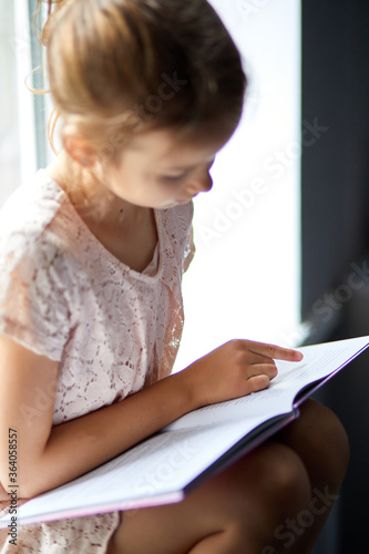 Cute little girl reading book at home, at windowsill