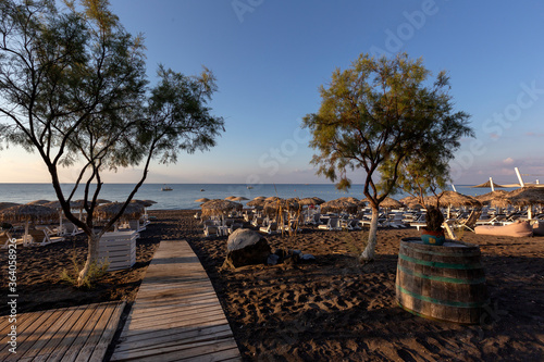 Santorini black beach Perissa, Perivolos illuminated by the morning rays of the sun. Beach chairs with umbrellas. Two olive trees. Beautiful calm sea.