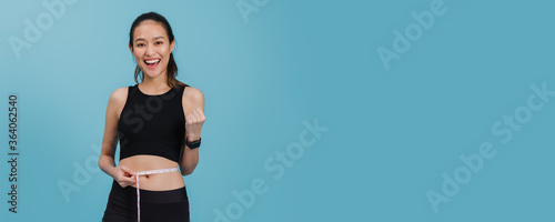 Portrait of confident beautiful asian fitness woman standing and use tape measure to measure waist  after exercise with smiling face on blue colour background.Concept of slim and healthy happy girl.