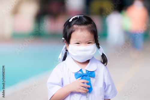 Sweet smiling schoolgirl is in the school area. Little child smiles at parents or mother. Wear a face mask in the situation disease Covid-19 pneumonia. Children wearing school uniforms aged 3 years.