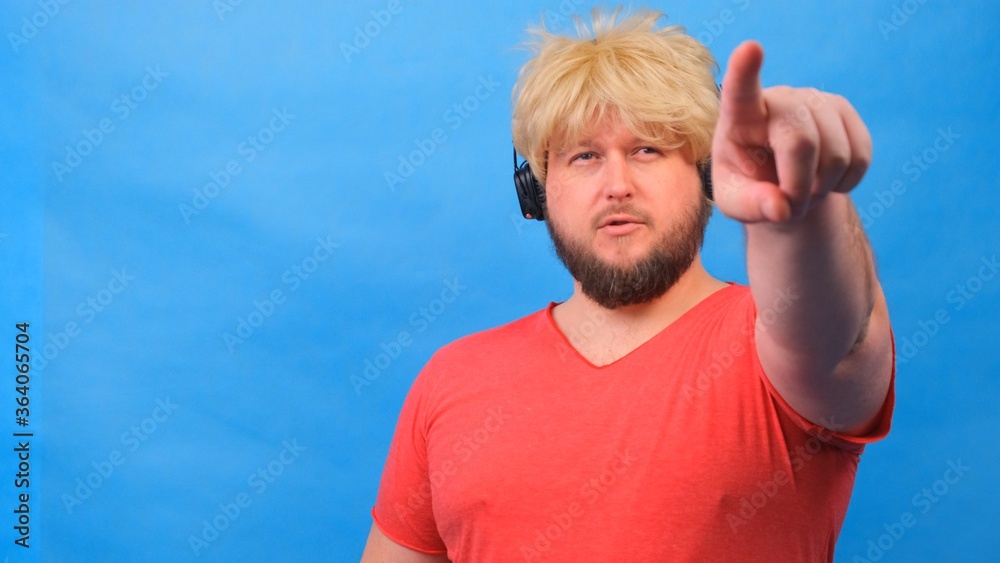 Naklejka premium freaky fat man in a wig and a pink T-shirt in headphones shows a finger on the camera at you against a blue background.