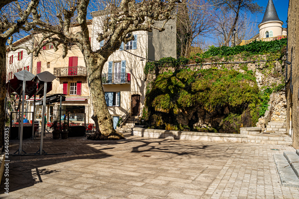 Provencal village in winter. Terrace of a bar in the sun. On the square, a mossy fountain and its basin. Plane tree with bare branches.