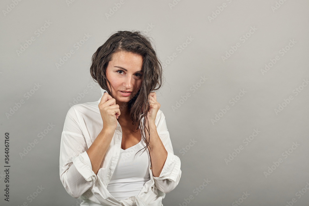 Teen smiling model brushing hand through hair, fashion posing, with hands in strand. Young attractive woman, dressed white blouse, with brown eyes, curly hair, gray background Teen smiling model brushing hand through hair, fashion posing, with hands in strand. Young attractive woman, dressed white blouse, with brown eyes, curly hair, gray background