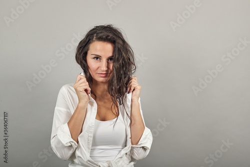Teen smiling model brushing hand through hair, fashion posing, with hands in strand. Young attractive woman, dressed white blouse, with brown eyes, curly hair, gray background