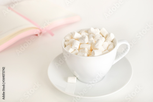 Tasty marshmallows in a mug with hot chocolate on a writing desk. Female desktop with a marshmallow mug and a notebook.