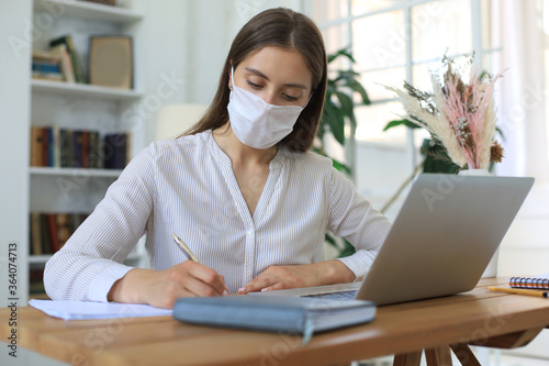 Young businesswoman in a medical protective mask works from home at the laptop during self-isolation and quarantine.