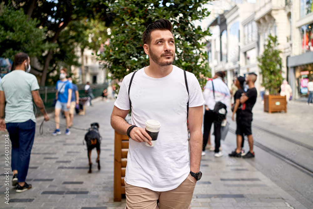 Obraz premium The man walking in the crowded street with his coffee in his hand is wearing a white T-shirt and canvas pants. man walking with his coffee in his hand on istiklal street