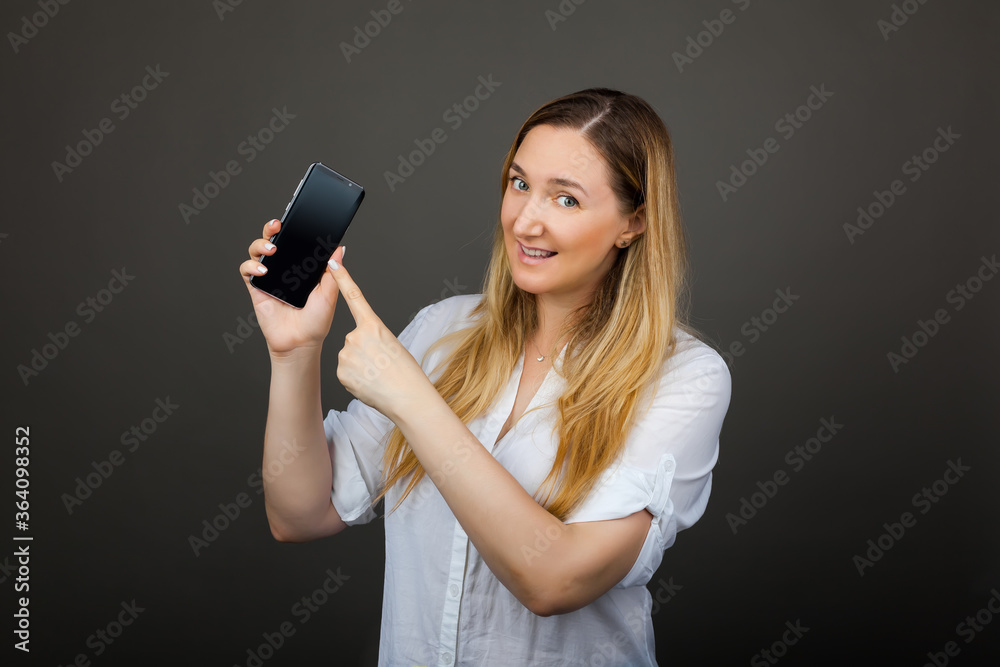 Smiling woman is pointing on smartphone standing on grey background. Smiling woman is pointing on smartphone standing on grey background.