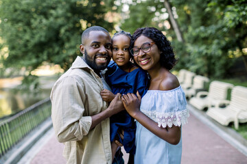 Smiling joyful family portrait in park. Portrait of young Arican family, standing together on park alley background, holding their little daughter in hands and laughing to camera