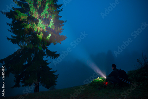 View of night tree, man with a flashlight in foggy mountain valley. Scenic landscape of misty hills under blue sky. Concept of travel and magical nighttime. photo