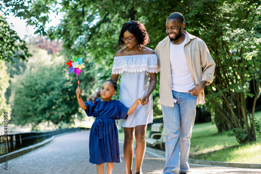 Happy family, joyful time together outdoors. Dark skinned little girl in blue dress, blowing colorful windmill toy, while walking in park alley with her young happy parents on summer day