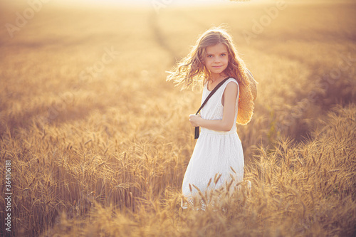 Fashion photo of a little girl in white dress and straw hat at the evening wheat field.