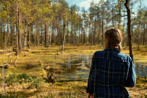 Rear view of unrecognizable young woman with ponytail having walk outdoors alone, posing in pine forest wearing coat, standing in front of swamp, enjoying beautiful sunny weather on spring day