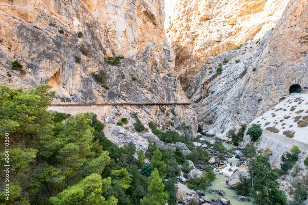 Mountain landscape view of El Chorro narrow gorge, river and El Caminito del Rey walkway wooden platforms attached to vertical rocks over the precipice, with tourists walking, Spain.