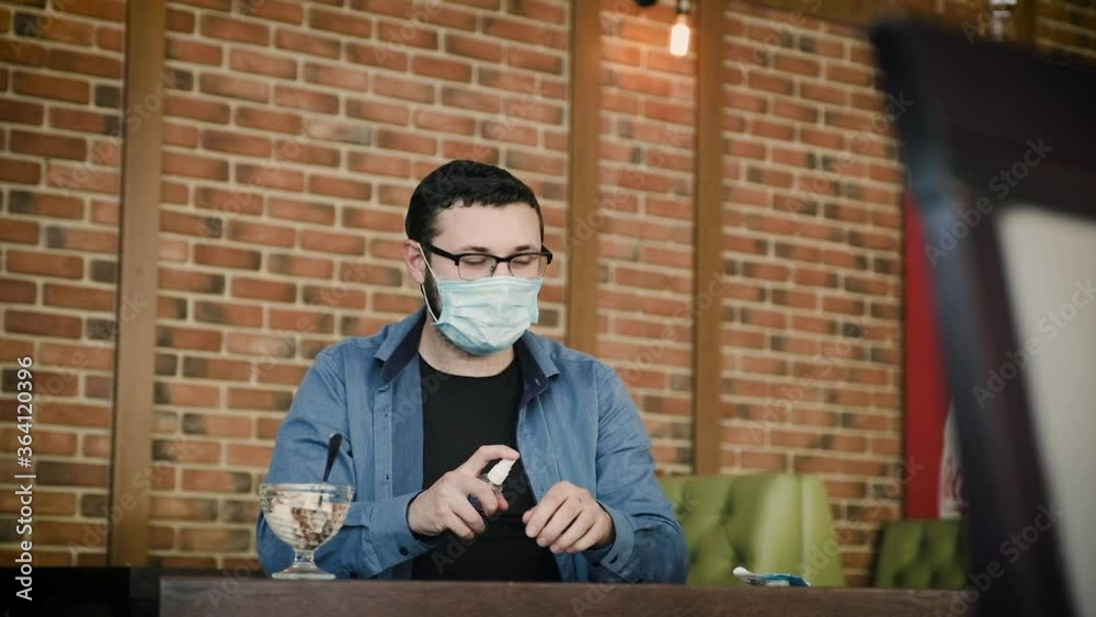 A young man uses hygiene products in a city cafe and without taking off his mask comically eats ice cream. A young man uses hygiene products in a city cafe and without taking off his mask comically eats ice cream.