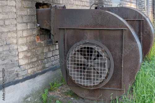 Industrial centrifugal radial exhaust fans of snail type of brown color with traces of rust, installed near building made of white silicate bricks with traces of soot, at  side of which grass grows