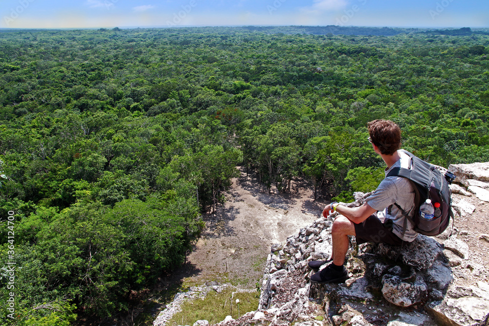 A young man with a grey backpack sitting on the top of the famous pyramid of Coba, Yucatan, Mexico. The jungle in the background reaches till the horizon. Concept of travel, nature and ancient culture