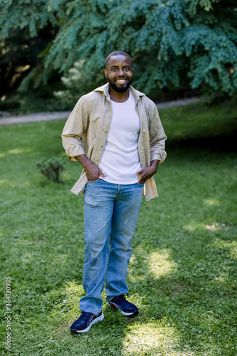 Summer outdoor portrait of handsome smiling african man in jeans, white t-shirt and beige casual shirt looking at camera while standing on green grass in park, enjoying the walk