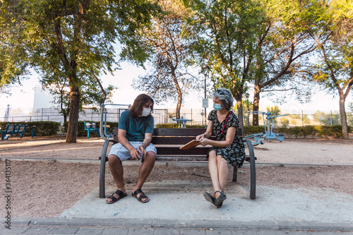 Young man and woman with mask consulting book sitting in urban park, keeping social distance to prevent the spread of the coronavirus in Spain. Selective focus.