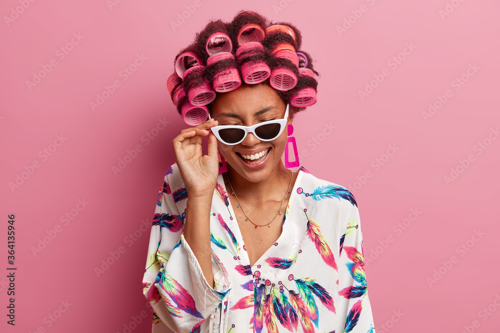 Overjoyed young woman has fun at home, laughs sincerely, wears sunglasses, hair curlers, dressed in dressing gown, hears funny story, isolated over pink background. Beauty and fashion concept