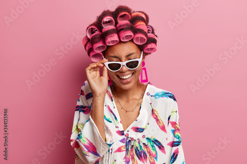 Overjoyed young woman has fun at home, laughs sincerely, wears sunglasses, hair curlers, dressed in dressing gown, hears funny story, isolated over pink background. Beauty and fashion concept