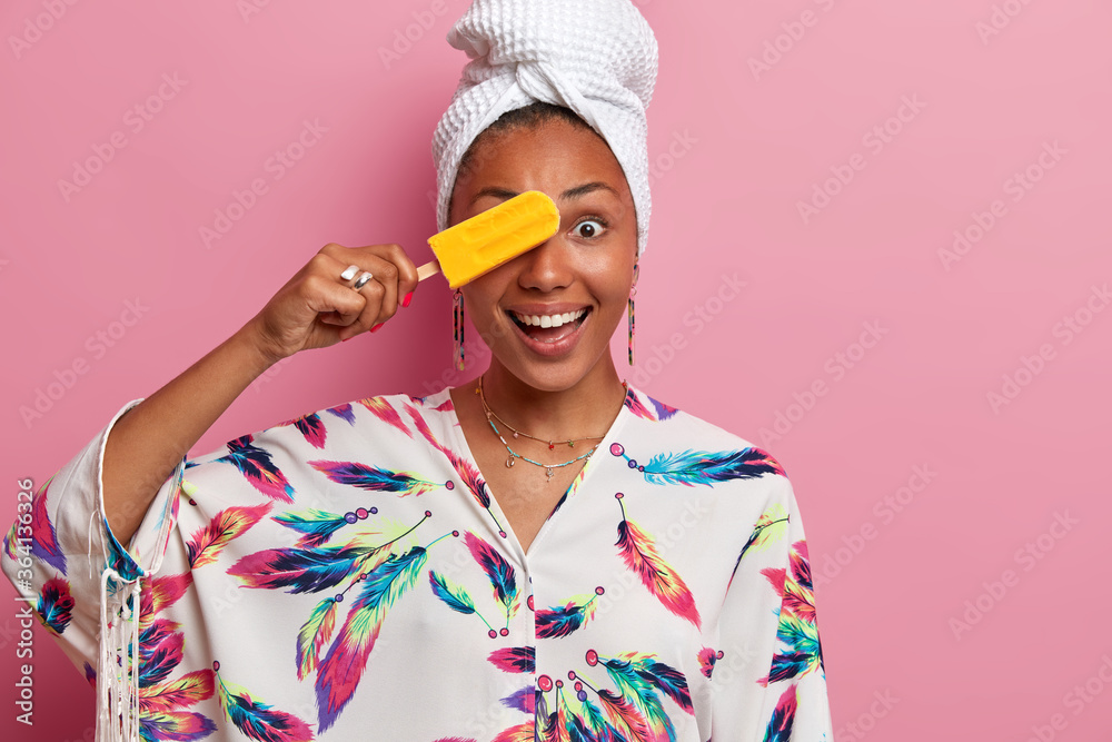 Domestic style and eating desserts. Smiling dark skinned young woman covers eye with refreshng yellow ice cream, has fun during summer time, wears dressing gown, poses against pink background Domestic style and eating desserts. Smiling dark skinned young woman covers eye with refreshng yellow ice cream, has fun during summer time, wears dressing gown, poses against pink background