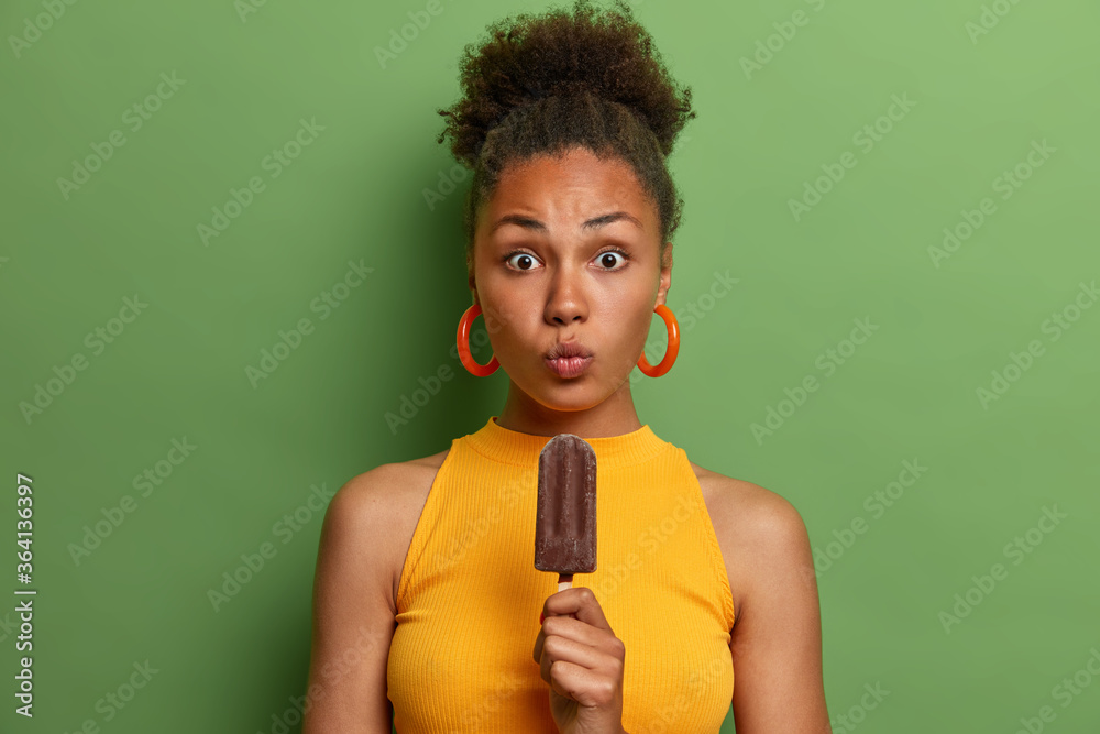 Surprised curly haired woman keeps lips rounded, eager to eat cold chocolate ice cream, spends weekend with friends during summer time, dressed in yellow clothing, poses over green vivid wall Surprised curly haired woman keeps lips rounded, eager to eat cold chocolate ice cream, spends weekend with friends during summer time, dressed in yellow clothing, poses over green vivid wall