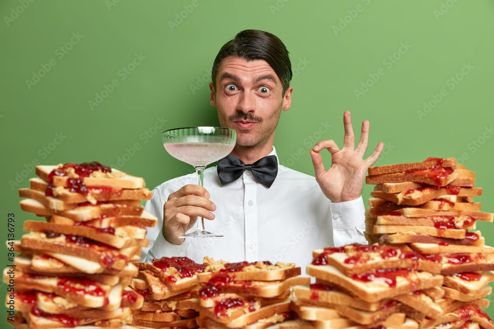 Handsome barman degustates cocktail, makes okay gesture, approval sign, says its tasty, dressed formally, poses against green background with bread toasts in foreground. People, food, drinking Handsome barman degustates cocktail, makes okay gesture, approval sign, says its tasty, dressed formally, poses against green background with bread toasts in foreground. People, food, drinking