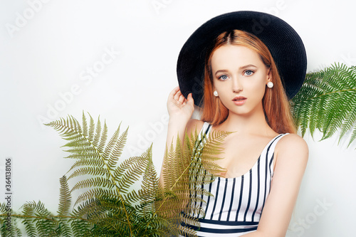 Girl in tropical leaves in a swimsuit in a big black hat smiles. Space for text, empty space. Model summer photo. Big earrings