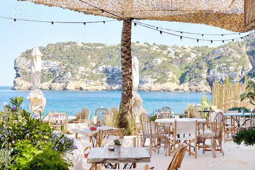 Fototapeta Naklejka Na Ścianę i Meble -  Interior of coastal beach restaurant. Under canopy located empty wooden tables and chairs, cozy cafe with beautiful view to rocky mountains hillside village and Mediterranean Sea bay. Sunny summer day