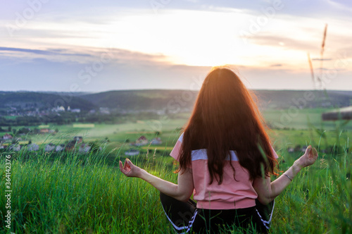 Photo of Young woman sitting back, meditate in lotus yoga pose outdoor, on the hill on the summer sunset