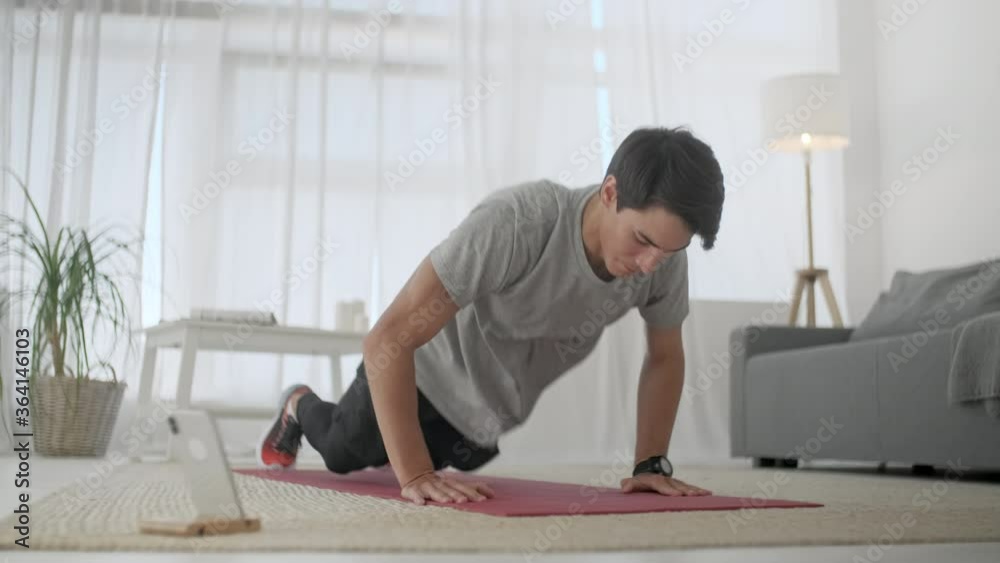 Handsome Young Man in a Gray T-Shirt and Black Shorts Does Sports at Home Online. Handsome Man Exercising and Doing Push ups in Living Room at Home. Handsome Young Man in a Gray T-Shirt and Black Shorts Does Sports at Home Online. Handsome Man Exercising and Doing Push ups in Living Room at Home.