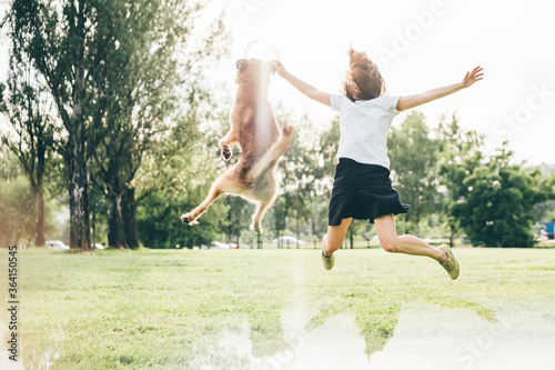 Pretty young lady with glasses in white t-shirt plays with jumping funny grey fluffy dog using plate on lush green lawn grass in sunny summer park