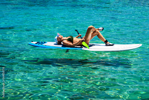Woman is lying on a SUP with some graffition, crystal clear waters and sunbathing on a hot sunny day. Healthy lifestyle concept. © Maksym