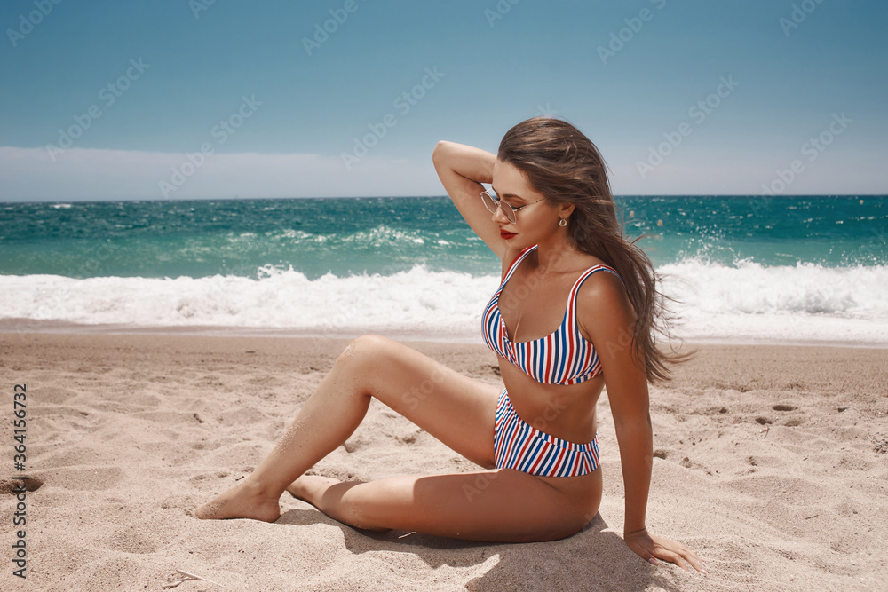 Sexy tanned caucasian woman in colorful striped bikini sitting at tropical beach against blue ocean on a sunny day. Beautiful girl with long dark hair. Fashion model posing outdoors