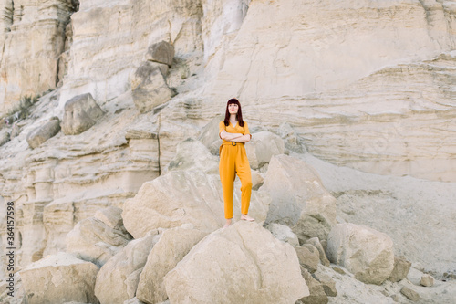 Full length horizontal summer portrait of a young brunette charming woman in fashionable yellow overalls, posing in sand quarry, standing on the stone. Summer fashion