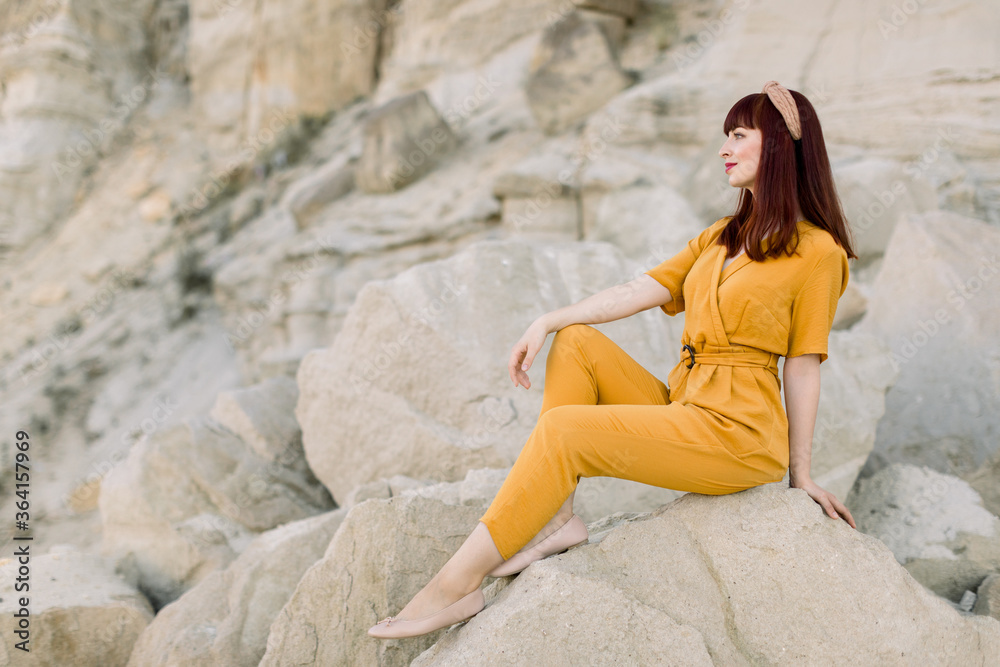 A young beautiful brown haired girl in fashionable yellow overalls poses on sunny summer day, sitting on the stone in front of sand quarry outdoors. Summer lifestyle and fashion concept. Copy space