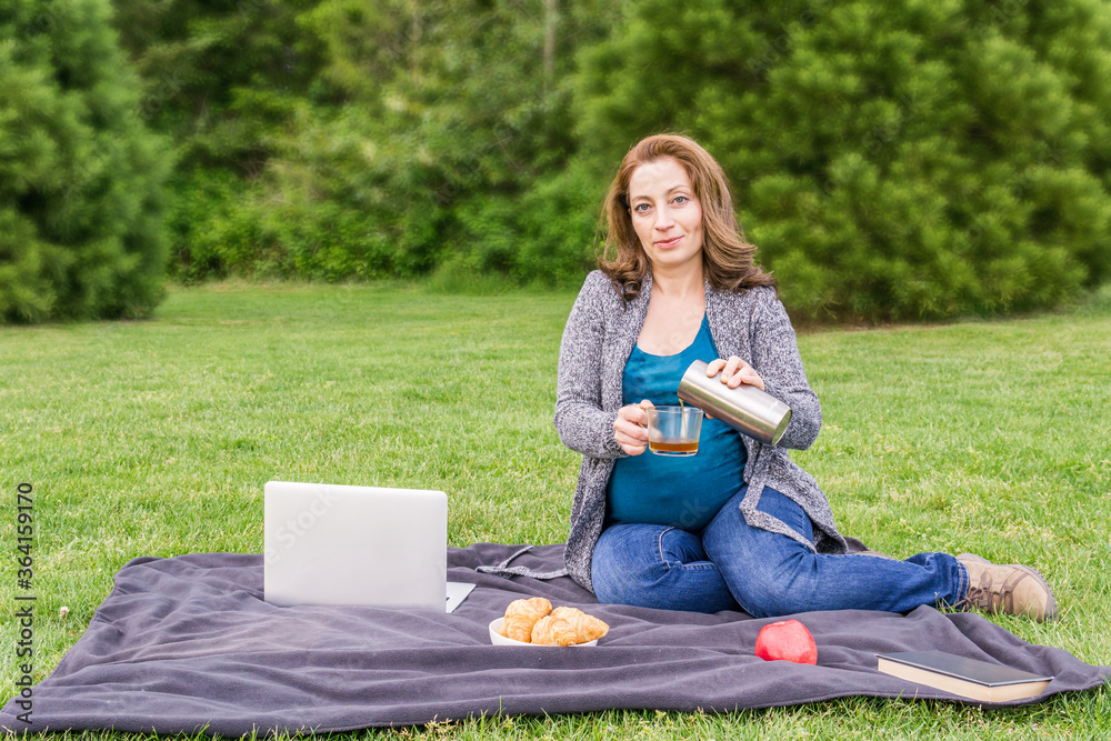 pregnant woman pouring coffee otr tea from stainless thermos in the park after working on a computer. pregnant woman pouring coffee otr tea from stainless thermos in the park after working on a computer.