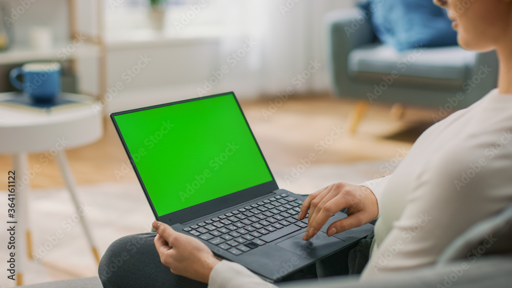 Young Woman at Home Sitting on a Couch Works on a Laptop Computer with Green Mock-up Screen. Girl Using Computer, Browsing through Internet, Watching Content, Chatting in Social Networks with Friends. Young Woman at Home Sitting on a Couch Works on a Laptop Computer with Green Mock-up Screen. Girl Using Computer, Browsing through Internet, Watching Content, Chatting in Social Networks with Friends.