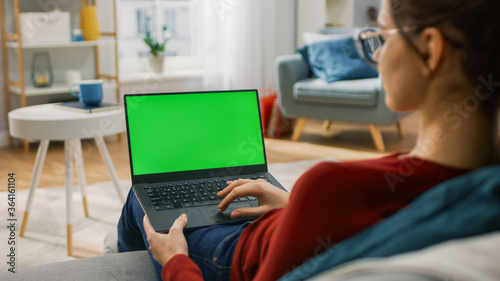 Young Woman at Home Works on a Laptop Computer with Green Mock-up Screen. She's Sitting On a Couch in His Cozy Living Room. Over the Shoulder Shot
