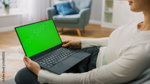 Young Woman at Home Sitting on a Couch Works on a Laptop Computer with Green Mock-up Screen. Girl Using Computer, Browsing through Internet, Watching Content, Chatting in Social Networks with Friends.