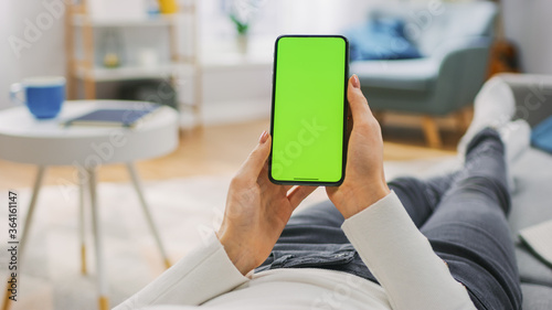 Young Woman at Home Laying on a Couch using with Green Mock-up Screen Smartphone. Girl Using Chroma Key Mobile Phone, Internet Browsing, Posting on Social Networks. Point of View Shot.