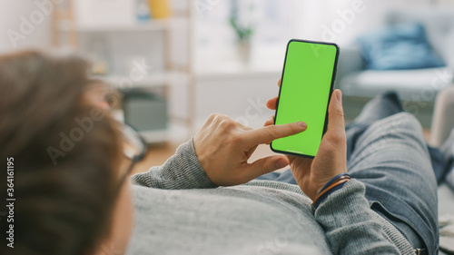 Man at Home Lying on a Couch using Smartphone with Green Mock-up Screen, Doing Swiping, Scrolling Gestures. Guy Using Mobile Phone, Internet Social Networks Browsing. 