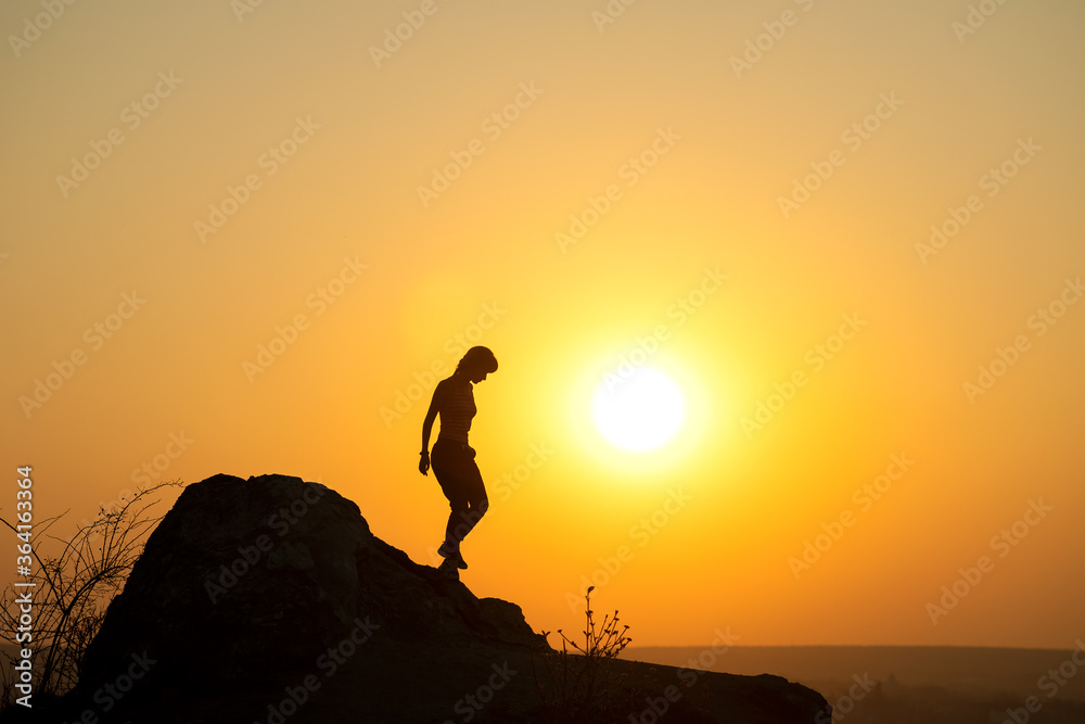 Silhouette of a woman hiker walking down a big stone at sunset in mountains. Female tourist on high rock in evening nature. Tourism, traveling and healthy lifestyle concept. Silhouette of a woman hiker walking down a big stone at sunset in mountains. Female tourist on high rock in evening nature. Tourism, traveling and healthy lifestyle concept.