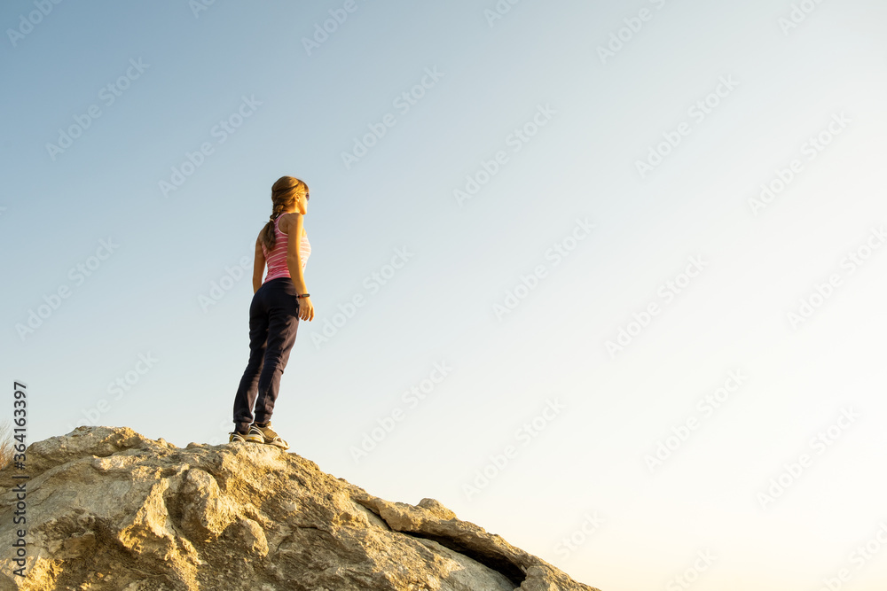 Young woman hiker standing alone on big stone in morning mountains. Female tourist on high rock in wild nature. Tourism, traveling and healthy lifestyle concept.
