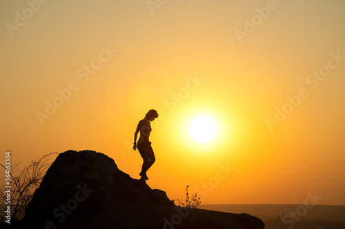 Silhouette of a woman hiker walking down a big stone at sunset in mountains. Female tourist on high rock in evening nature. Tourism, traveling and healthy lifestyle concept.